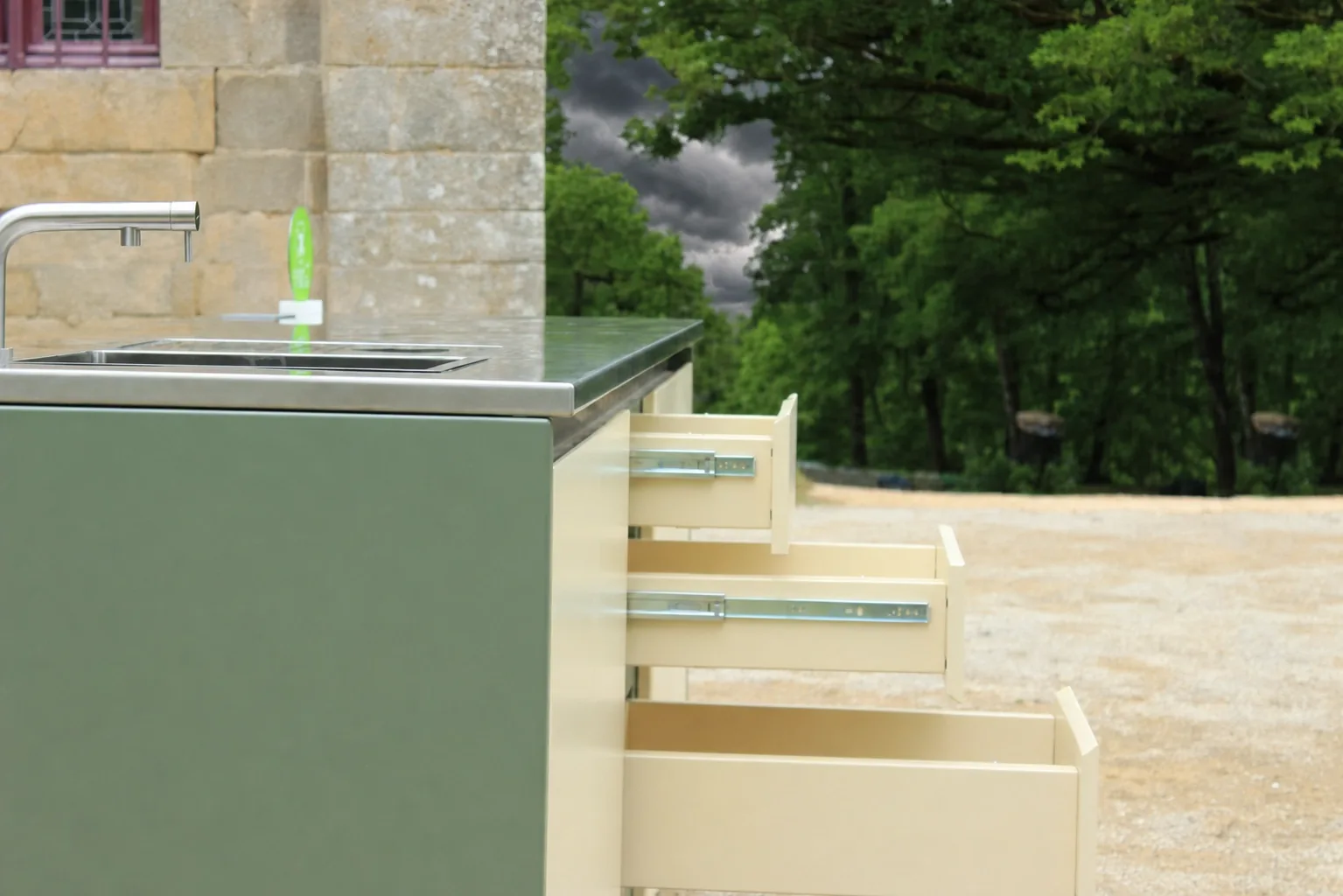Tiroirs à extension totale ouverts sur la cuisine d'extérieur mobile ROLYX, plan de travail en inox et évier intégré, sur le gravier d'une cour