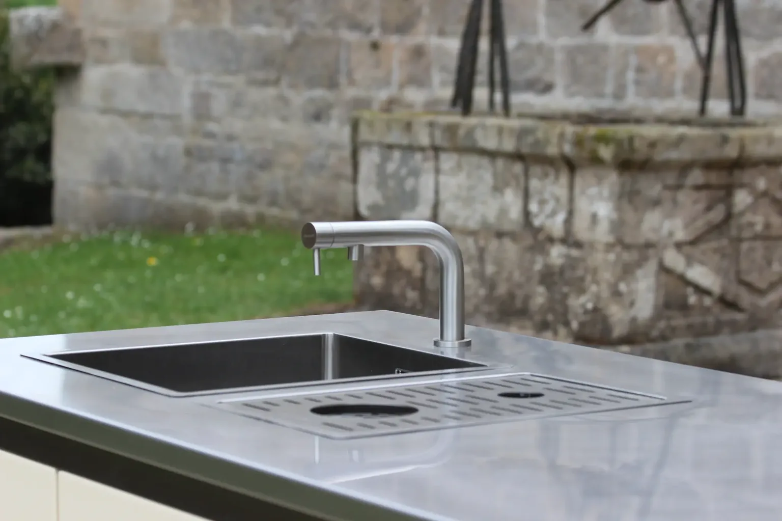 Évier et rince-verres en inox intégrés au plan de travail d'une cuisine d'extérieur ROLYX, devant un mur de pierre