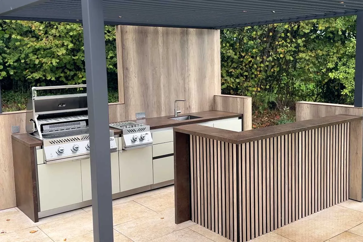 Cuisine d'extérieur SOLSTYS installée sous une pergola bioclimatique, barbecue et brûleur encastrés, bar en tasseaux de bois