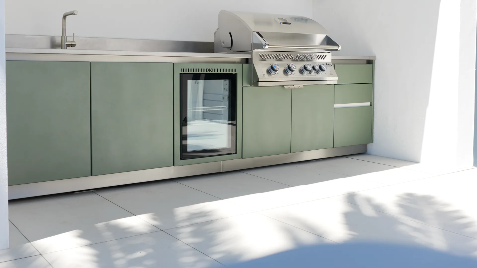 Cuisine extérieure EKZALT aux façades vert sauge : évier, réfrigérateur vitré et barbecue encastré sur un plan en inox, entre deux murs