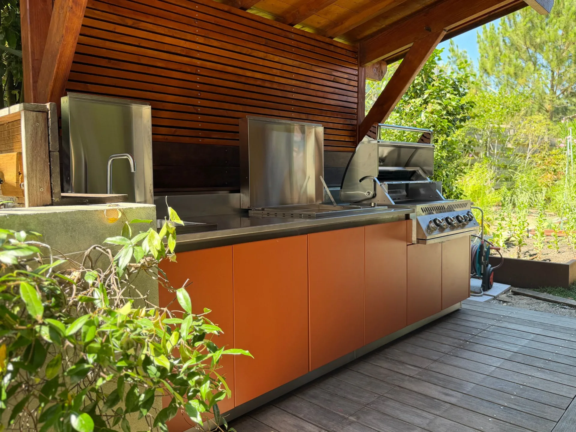 La cuisine EKZALT orange sous sa pergola en bois, vue de biais, la terrasse et le jardin derrière