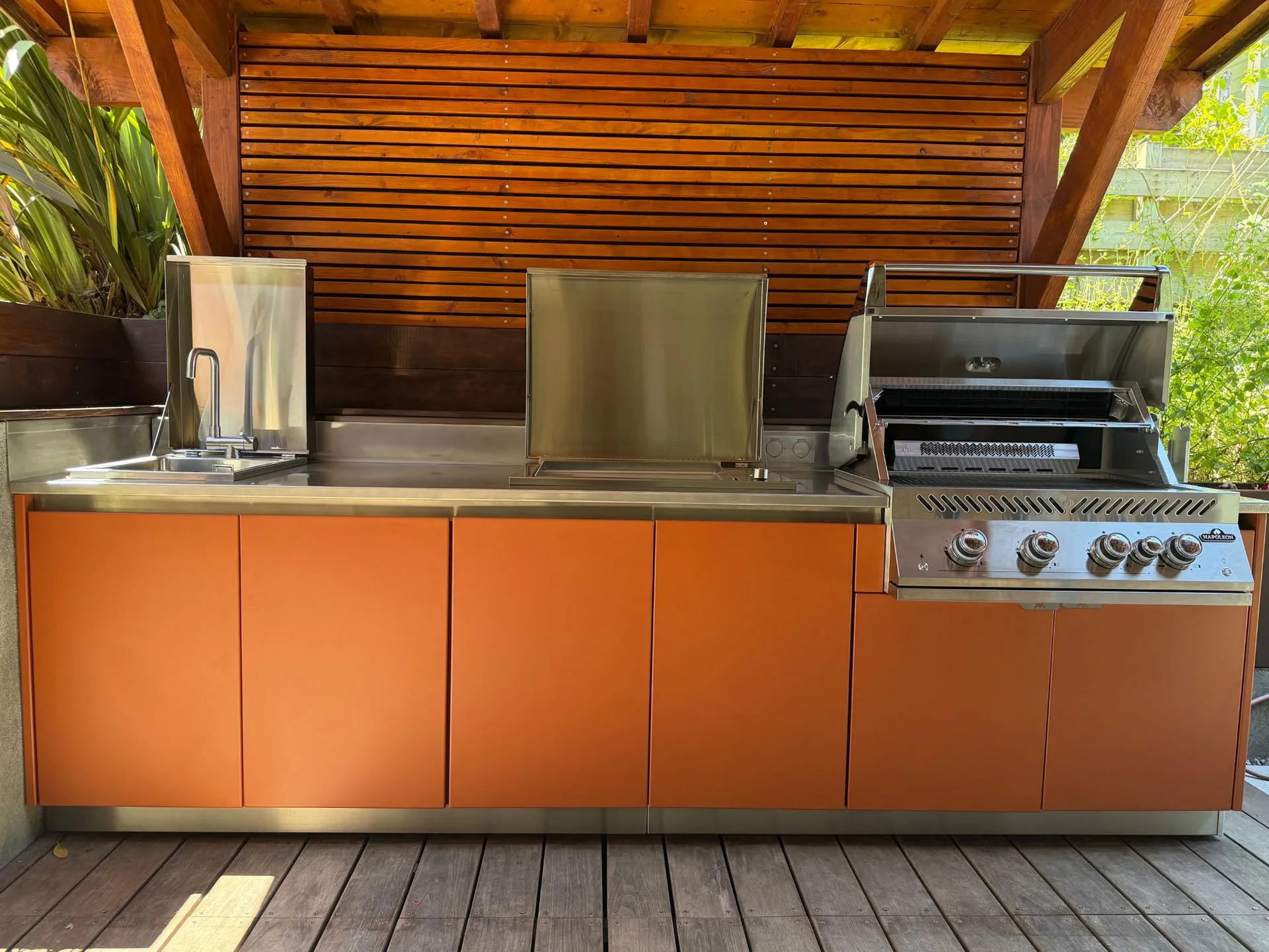 Cuisine extérieure EKZALT aux façades orange sous une pergola en bois, vue de face : évier, plancha et barbecue, capots relevés