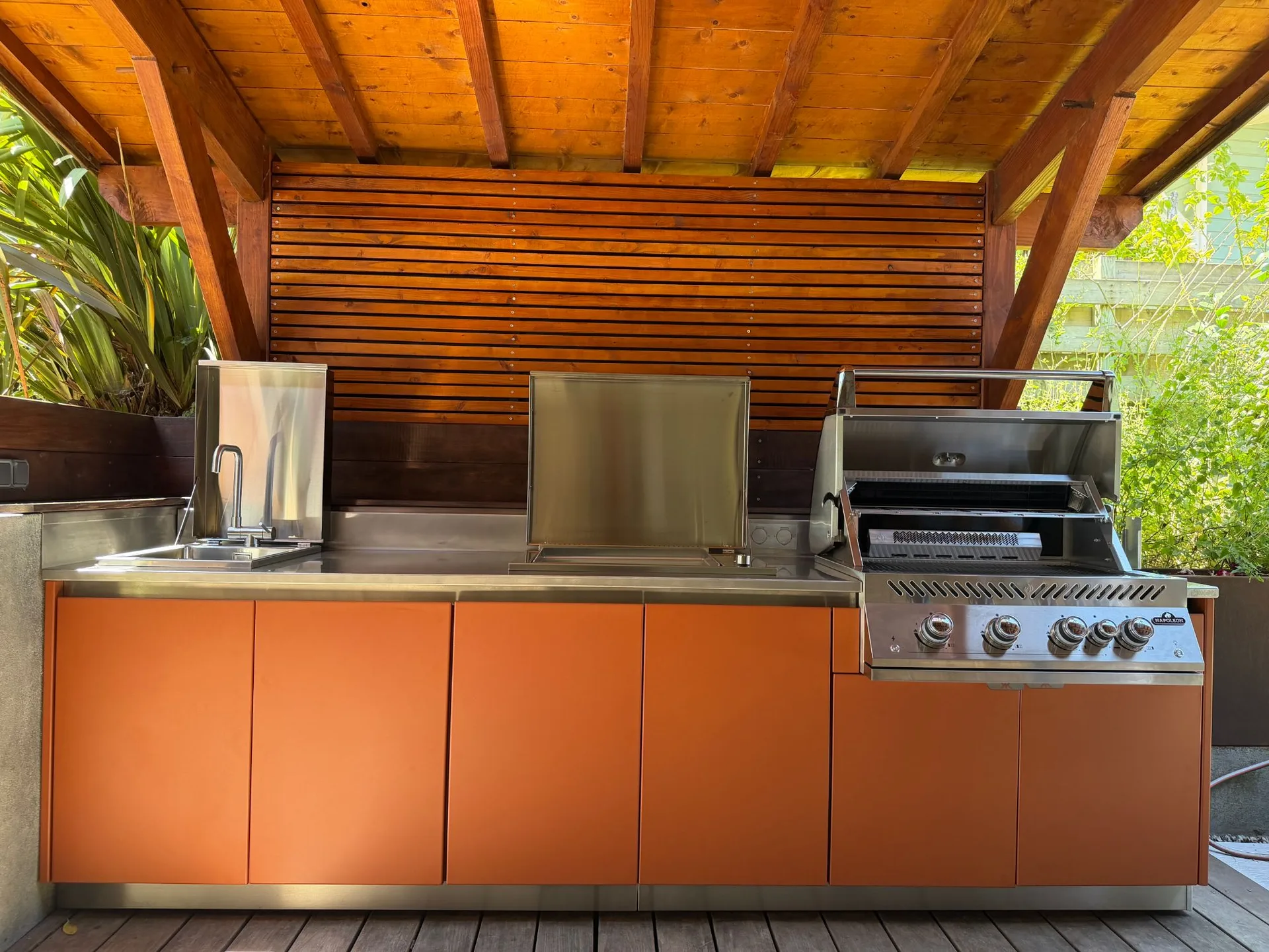 Les façades orange de la cuisine EKZALT sous le toit en lames de bois de la pergola