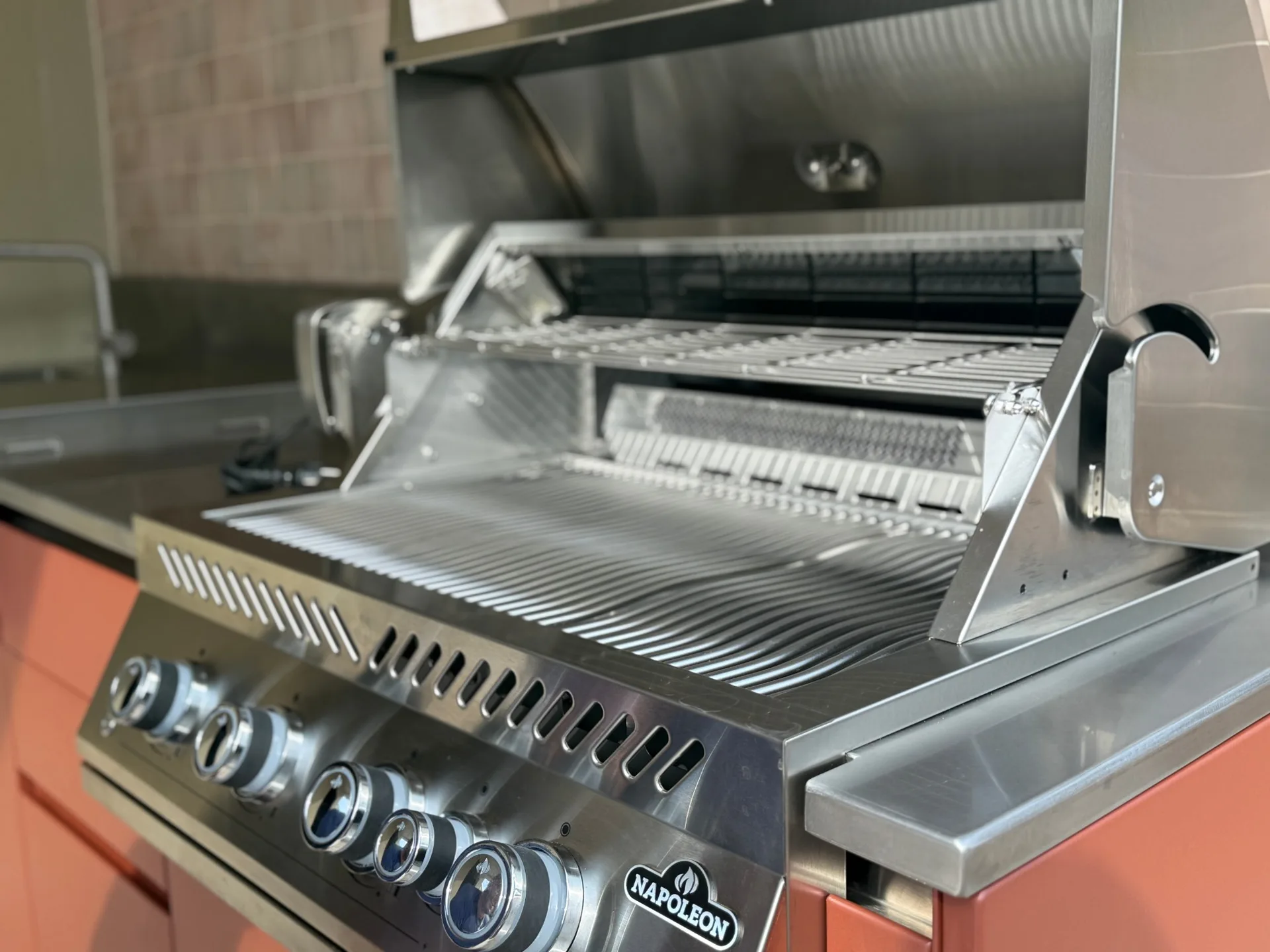 Le barbecue Napoleon capot ouvert, grilles en vagues et brûleur arrière, encastré dans le plan inox de la cuisine EKZALT aux façades Lahoré