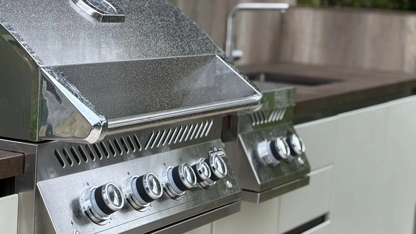 Barbecue et brûleur latéral encastrés dans le plan de travail d'une cuisine d'extérieur EKINOX, façades claires et mitigeur en arrière-plan