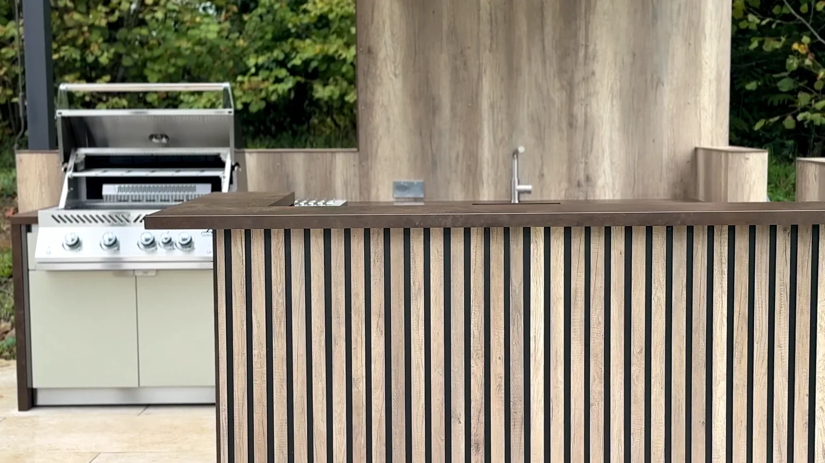 Le bar en tasseaux de bois de la cuisine SOLSTYS de Pompadour, le barbecue Napoleon capot ouvert et l'évier derrière, devant un panneau façon bois
