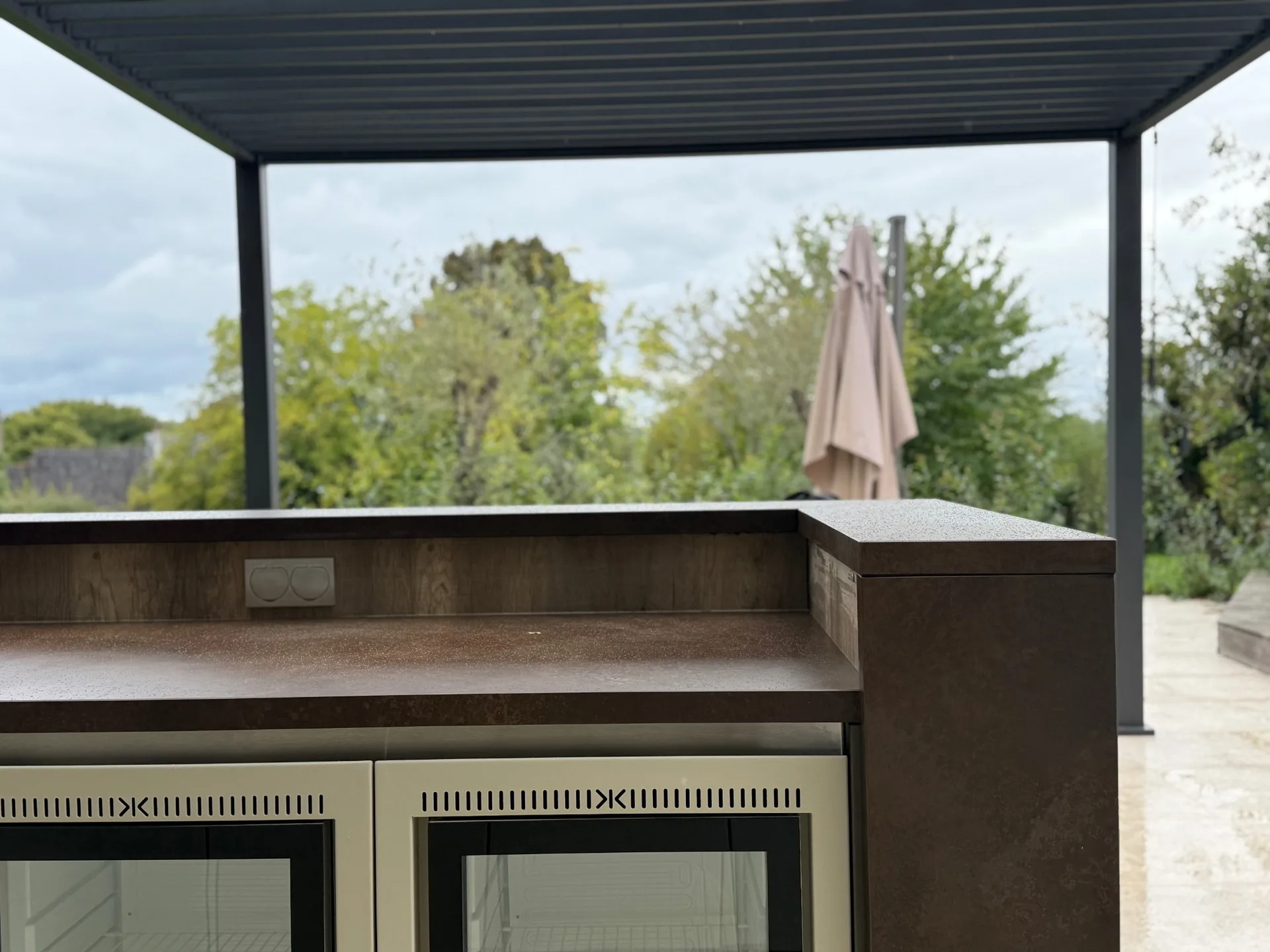 Le comptoir du bar en céramique brune sous la pergola bioclimatique, les deux réfrigérateurs vitrés et les prises étanches, le jardin derrière