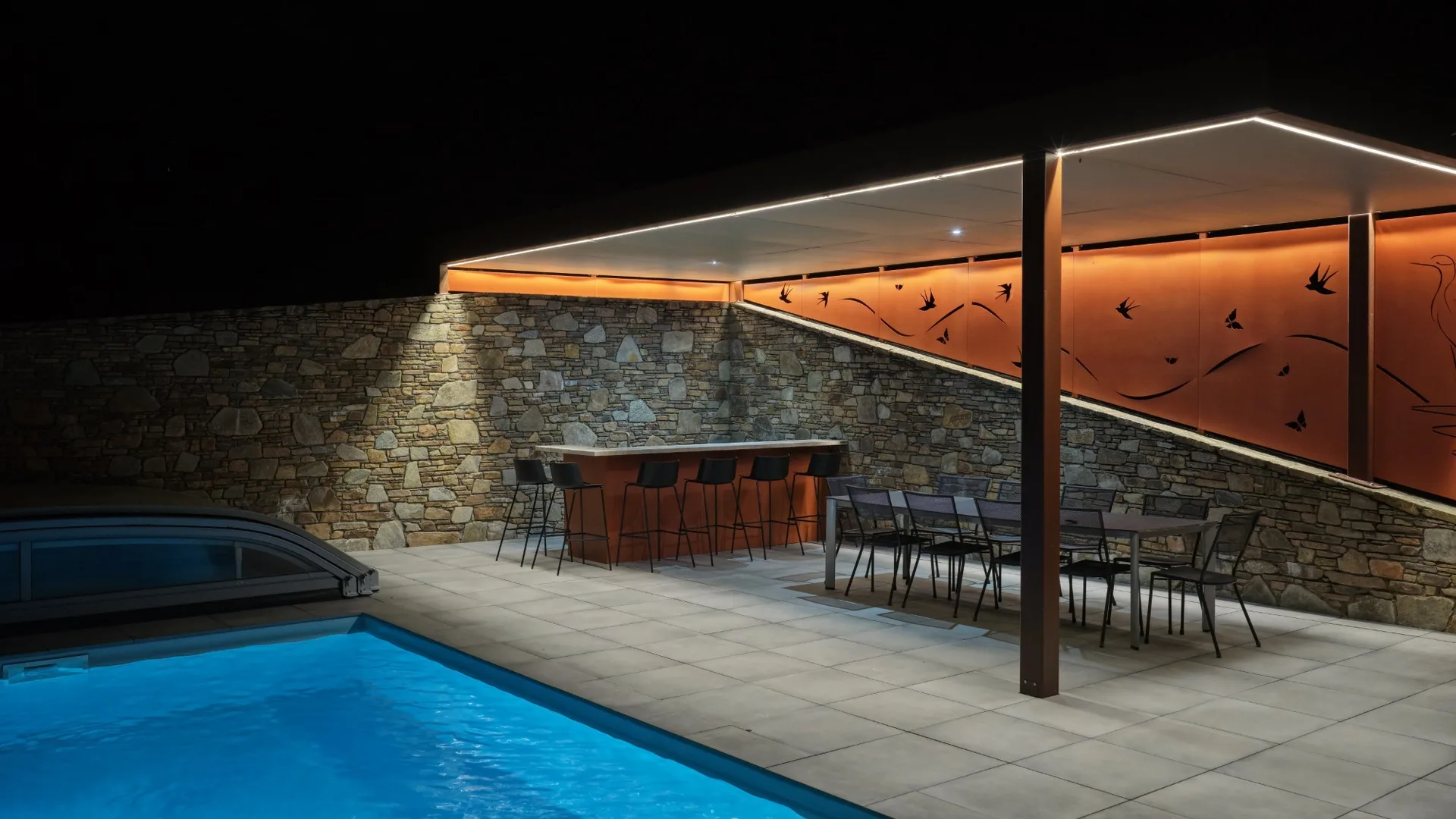 Bar de cuisine extérieure SOLSTYS éclairé sous sa pergola, de nuit, vu depuis la table de la terrasse
