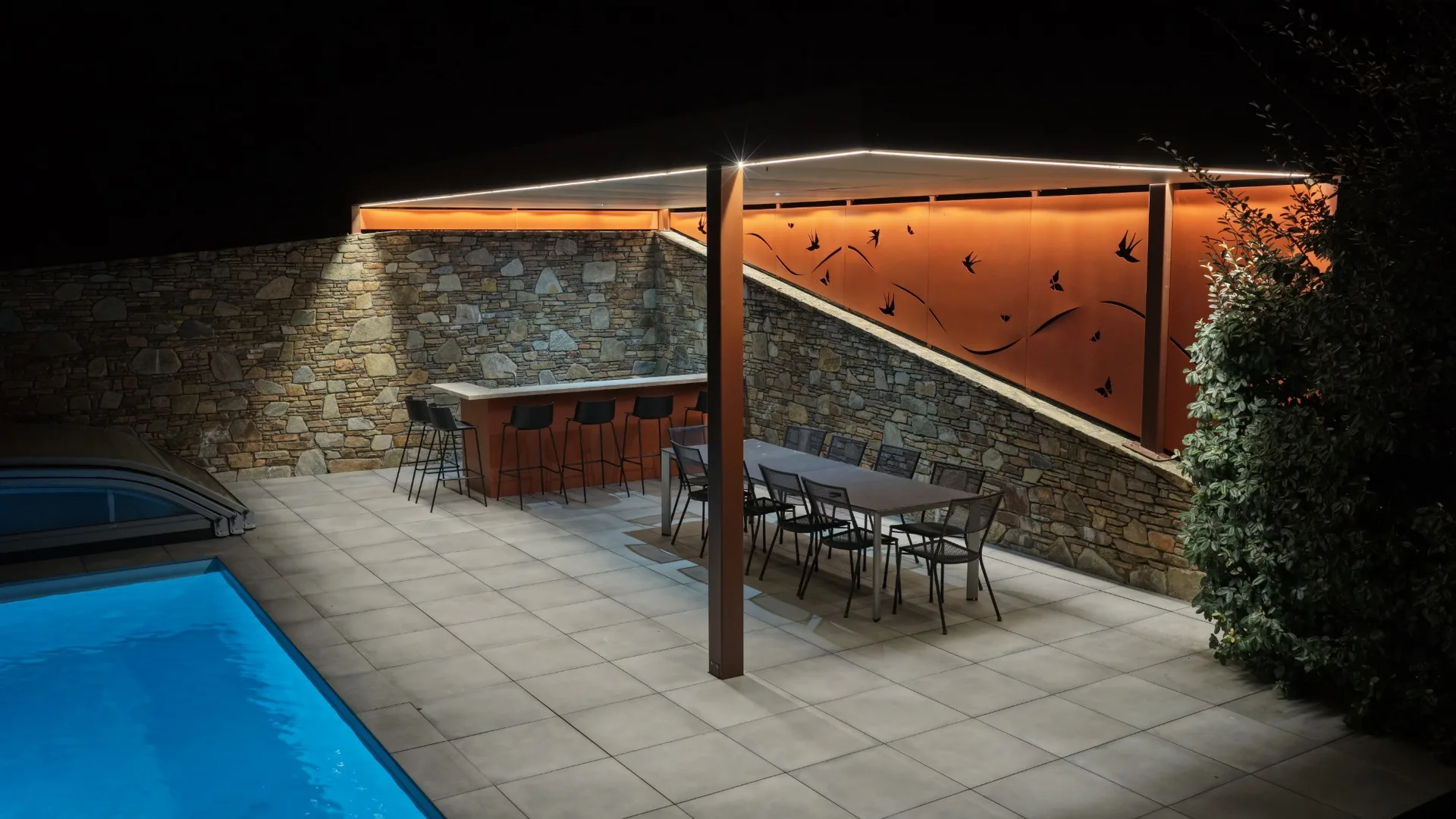 Le bar SOLSTYS et sa pergola éclairés, vus de nuit depuis le bord de la piscine