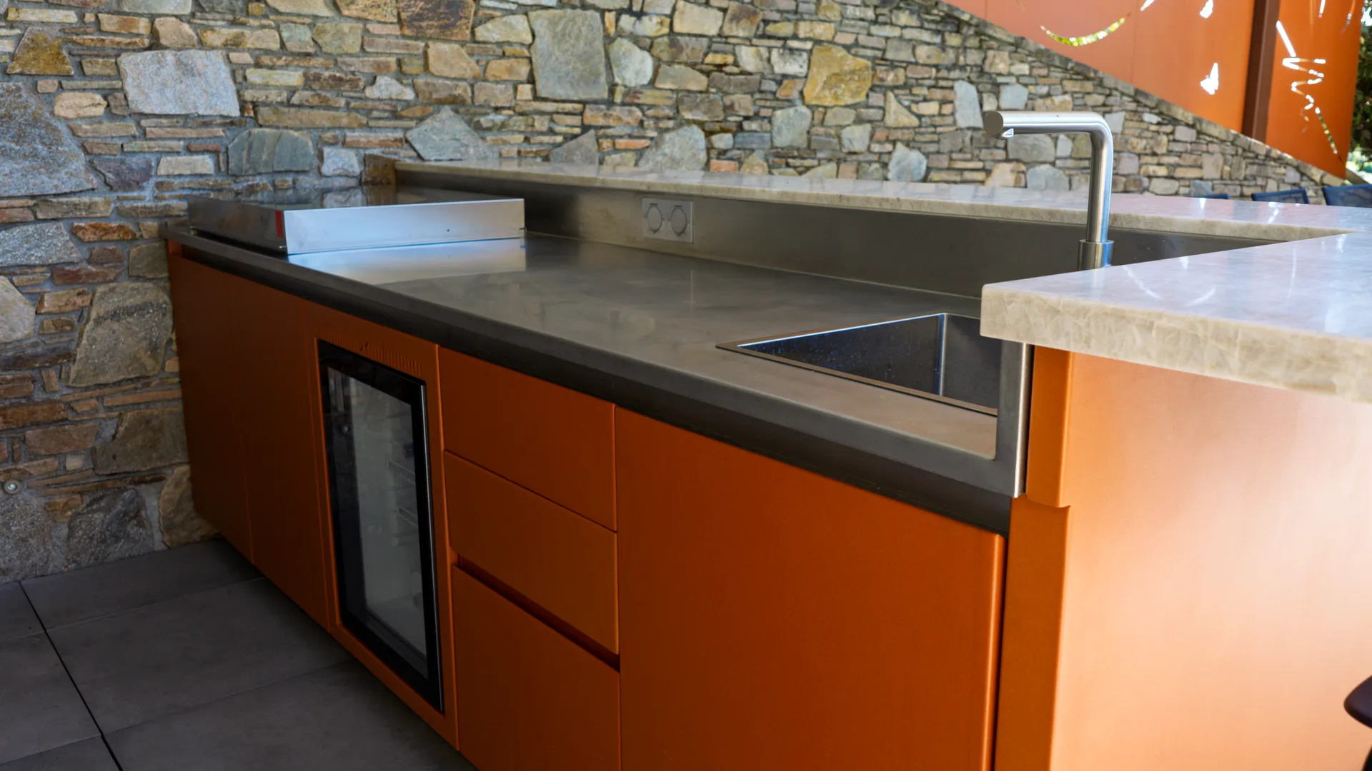 Le plan en inox 316L et les façades orange du bar SOLSTYS, de jour, le mur de pierre derrière