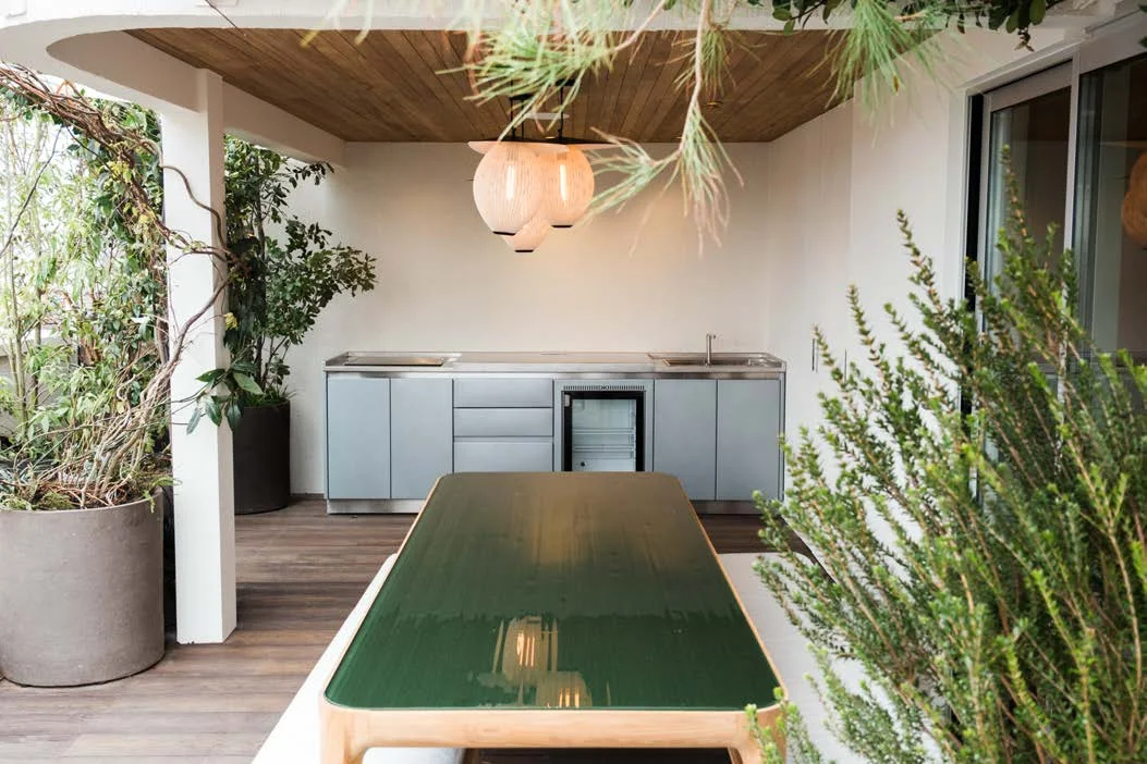 Cuisine d'extérieur SOLSTYS en ligne sous l'auvent d'un rooftop parisien, plan de travail inox, réfrigérateur encastré et grande table de repas