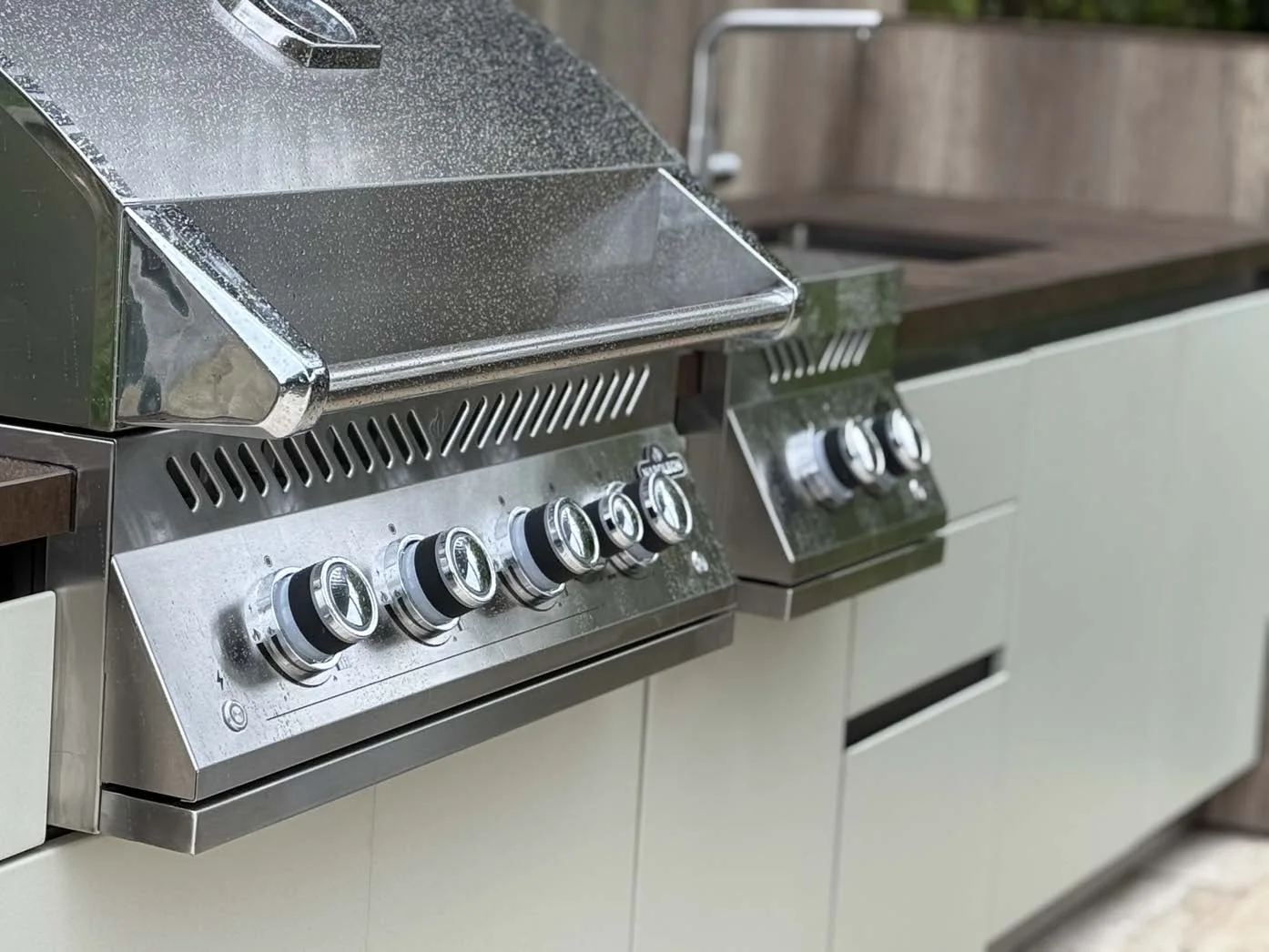 Barbecue encastré capot relevé et brûleur latéral dans un plan de travail en céramique brune, façades gris blanc, évier au second plan