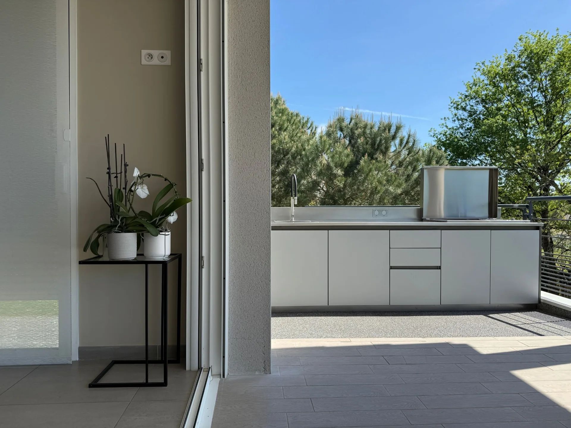 La cuisine X-PRESS vue depuis le salon, porte-fenêtre ouverte : plan inox, évier, plancha sous son couvercle, les pins derrière la terrasse